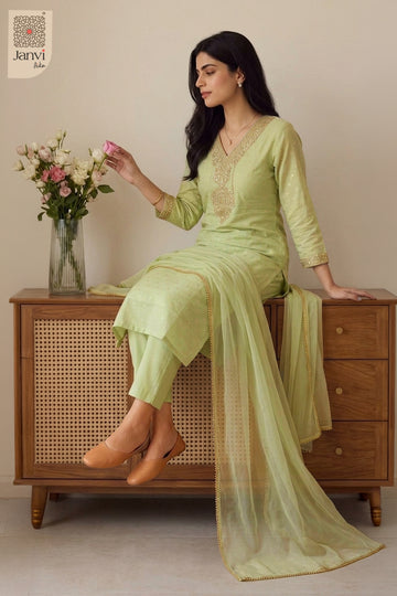 Seated pose beside a floral vase wearing an elegant pastel sage silk churidar set.